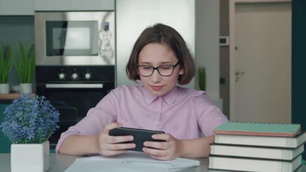Little Girl with glasses Sitting into the desk Using Smartphone Watching Video Child is bored Technology at home. Quarantine concept. Distance education. How to keep your child busy. — Vídeo de Stock