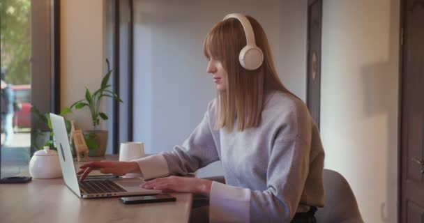 Joven y hermosa mujer rubia en los auriculares sentado en la cafetería está trabajando en el ordenador portátil. Feliz freelance o empleado navegando por Internet y charlando con amigos, tomando café. — Vídeo de stock