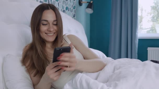 Een jonge vrouw met donker haar typt berichten naar een vriendje op de smartphone nadat ze wakker werd in de slaapkamer. Een vrouw is blij met berichten op haar mobiele telefoon — Stockvideo