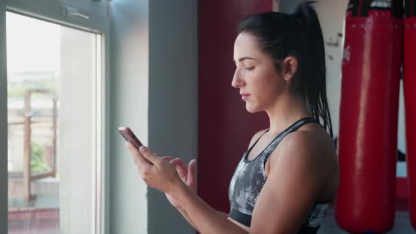 Vrouw met smartphone met personal trainer workout app in de sportschool. Vrouw bereidt zich voor op de training met behulp van fitness app staan in de buurt van raam. — Stockvideo