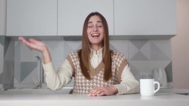 Çelik yelek giyen çekici, yetişkin bir kadın vlogger kamera karşısında canlı görüntülü konferans, blog kaydı, modern mutfak arka planında bir sandalyeye oturuyor.