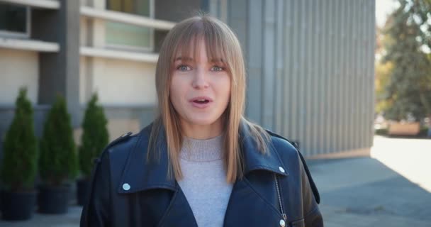 En chockad blond kvinna i svart läder jacka säger wow och täcker hennes mun när hon står urban stad bakgrund. — Stockvideo