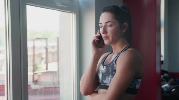 Retrato de una joven gurú de la aptitud física de cabello atado en una cola de caballo, en un traje para la aptitud hablando en el teléfono inteligente de pie en el gimnasio cerca de la ventana. Mujer llamando a clientes para decidir problemas de trabajo — Vídeos de Stock