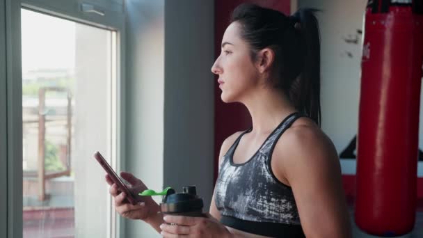 Νεαρή γυναίκα στον αθλητισμό στολή στέκεται κοντά στο παράθυρο serving ειδήσεις σε smartphone και ποτά πολυβιταμινούχο ποτό το πρωί, τη φροντίδα της υγείας του, απολαμβάνοντας ηλιόλουστο καιρό στο γυμναστήριο. — Αρχείο Βίντεο