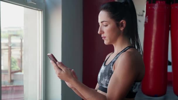 Feminino artes marciais masterusing telefone inteligente com personal trainer aplicativo de treino no ginásio. Mulher se preparando para o treino usando o aplicativo de fitness de pé perto da janela. — Vídeo de Stock