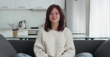 Modern dairede rahat bir koltukta oturan ve video konferansına katılanları dinleyen bir kadın. Başını salla ve kameraya gülümse..