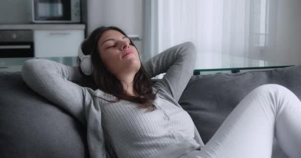 Ηρεμία νεαρή γυναίκα σε ακουστικά έχοντας υγιή ύπνο την ημέρα χαλαρώνοντας στον καναπέ με τα μάτια κλειστά χέρια πίσω από το κεφάλι, ήσυχο κορίτσι κοιμάται αναπνοή φρέσκου αέρα στηρίζεται σε άνετο καναπέ του — Αρχείο Βίντεο
