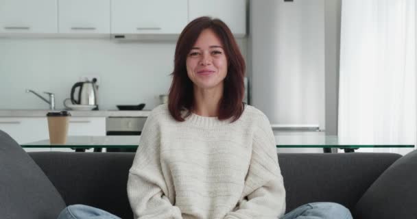 Woman sitting in cozy sofa in modern apartment and listens to the participants of the video conference . Nodding your head and smiling looks at the camera. — Wideo stockowe