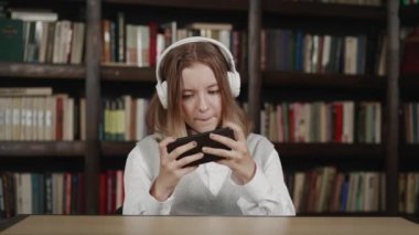 Beyaz kulaklıklı tembel liseli kız ev ödevlerini yapmaktan alıkoyan akıllı telefonlu kütüphanede oturuyor.
