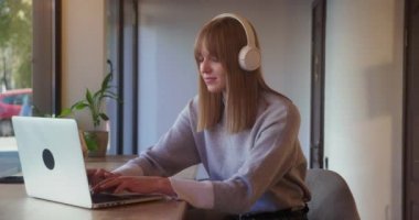 blonde woman in white headphones typing message on laptop sitting in cafe. Freelance concept, business correspondence.