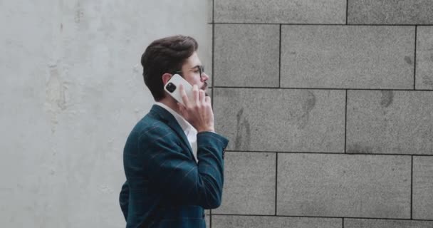 現代的な建物に沿って歩いている間、ビジネスマンは電話で話しています。コンセプトビジネス会話. — ストック動画