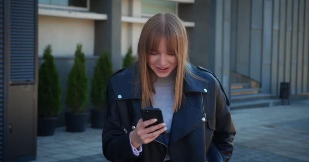 Blonde young woman in a leather jacket uses her phone while standing in the city centre. Urban buildings background. — Wideo stockowe
