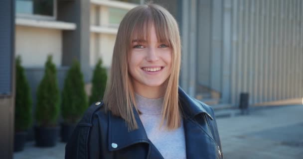 Retrato de una hermosa mujer rubia con el pelo sonriendo encantadoramente mientras está de pie en el paisaje urbano de la ciudad, con chaqueta de primavera Happy Young Woman disfruta de la vida. — Vídeos de Stock