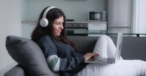 ソファのリビングルームにノートパソコンで座ってる若い大人の女性。ヘッドフォンを持つ女性は、コンピュータ上のインターネット上の自宅から仕事や研究 — ストック動画