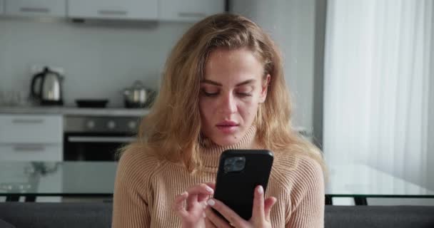 Curly blonde woman looking in smartphone typing message or surfing news sitting in the cozy sofa. Relax concept, free time or leisure — Stockvideo
