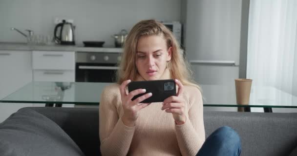 Jonge blonde haarvrouw die graag telefoneert. Voorraadbeelden. Stijlvolle jonge vrouw leuk en enthousiast spelen van games op uw smartphone zitten in de woonkamer op keuken achtergrond. Winnen — Stockvideo