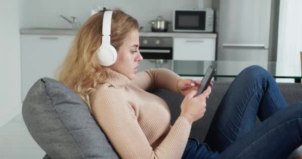 Détendu belle jeune femme jouissant de la musique avec smartphone profiter week-end tout en étant couché sur le canapé à la maison. — Video