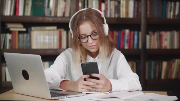 Surprised young teen girl with glasses sit of the table at library holding phone looking at screen watching social media video content, movie or stream online in mobile app laughing using smart phone. — Wideo stockowe