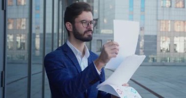 İş adamı belgeleri inceliyor ve düzenlemeler yapıyor. Kulaklık takıyor. Takım elbise ve gözlük takıyor. Arka planda iş merkezi.