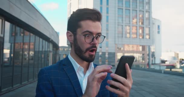 Ενθουσιασμένος νεαρός επιχειρηματίας με επίσημο κοστούμι και γυαλιά κοιτάζοντας στο smartphone δείχνει ναι χειρονομία. Ευτυχισμένος αρσενικός νικητής γιορτάζει την επιτυχία σε υπαίθριο κοντά σε ουρανοξύστες. — Αρχείο Βίντεο