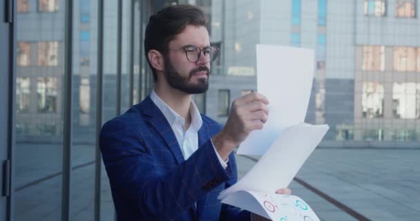 The businessman is looking through the documents and making edits. Hes wearing headphones. Hes wearing a suit and glasses. Business center in the background. — Stockvideo