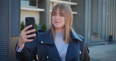 attractive blonde woman in black leather jacket talking by videocall on smartphone and waving at camera in urban city background.