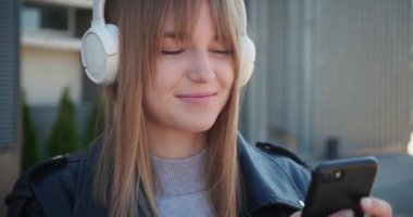 Beyaz kulaklıklı mutlu sarışın kadın akıllı telefondan müzik dinliyor ve dışarıda ayakta gülümsüyor. Kapatın. Boş zaman kavramı.