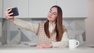 Güzel bir kadın cep telefonuyla selfie çekiyor mutfak arka planındaki masada oturuyor..