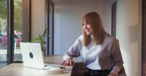 Młoda towarzyska blondynka radośnie przemawia video czatem na laptopie siedząc w przytulnej kawiarni — Wideo stockowe