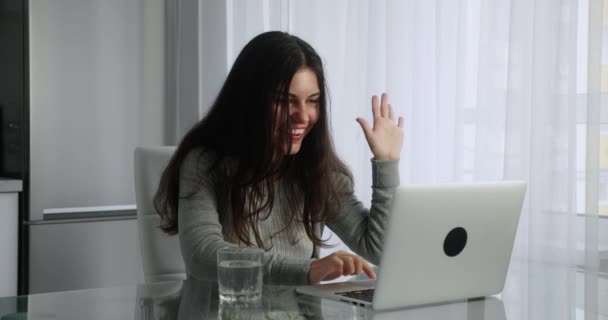 Mujer morena joven con el pelo largo tienen video chat en su computadora portátil sentado de fondo interior de la cocina. Una mujer sonríe agitando la mano mientras mira la pantalla — Vídeos de Stock