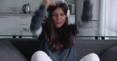 Oyun çubuğuyla video oyunu oynayan uzun saçlı komik çekici kız. Modern mutfak arka planında gri koltukta oturuyor. Kazanan konsept