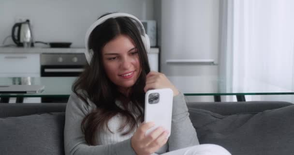 Entspannend. Glückliche Frau mit Kopfhörern, die Videos anschaut, Nachricht auf dem Handy tippt und auf der Couch sitzt — Stockvideo