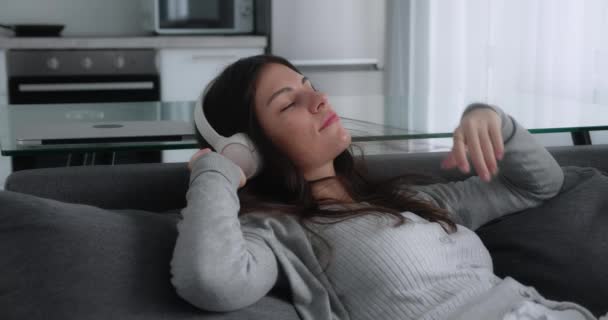 Tired young girl leans on the back of the sofa and listens to music on headphones while sitting on the sofa against the backdrop of the kitchen — Vídeo de Stock