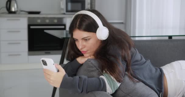 Teenage girl listening to music wearing headphones relaxing on sofa texting using smartphone browsing online playing mobile games enjoying sharing messages on social media — Stockvideo