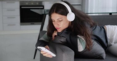 teenage brunette girl listening to music wearing headphones relaxing on sofa texting using smartphone browsing online playing mobile games enjoying sharing messages on social media