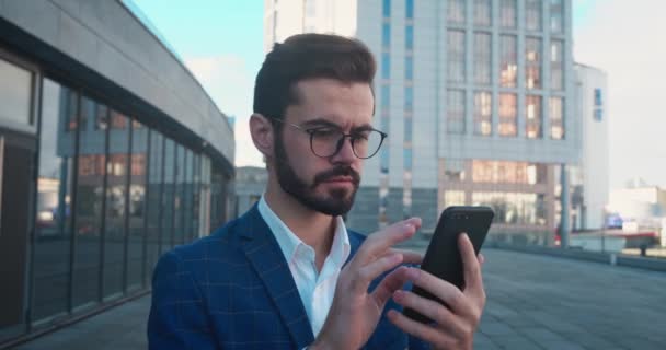Ευτυχισμένος έκπληκτος επιχειρηματίας κοιτάζοντας smartphone και λέγοντας WOW. Ευχάριστα έκπληκτος νεαρό αρσενικό πορτρέτο συναίσθημα σε υπαίθριο κοντά σε γυάλινο κτίριο. — Αρχείο Βίντεο