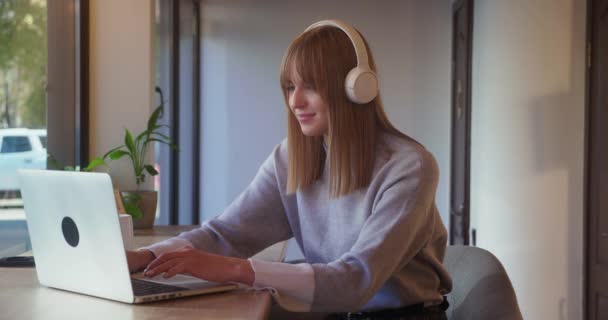 Young blonde woman freelance is working with laptop in cafe typing smiling sitting at table alone with portable computer. People and modern technology concept — стоковое видео