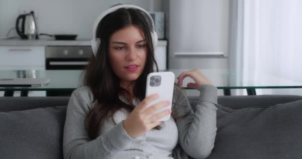A shocked brunette girl in a grey sweater saying wow looking smartphone and covering her mouth while sitting in couch kitchen background — Stockvideo
