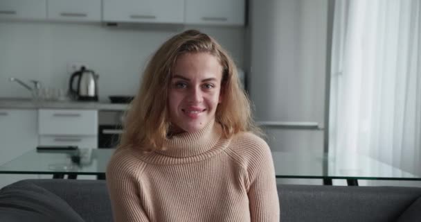 Ren lykke. Lykkelig positiv blondine krøllet kvinde i en varm sweater griner på kameraet, sidder i sofaen derhjemme, close-up portræt. – Stock-video