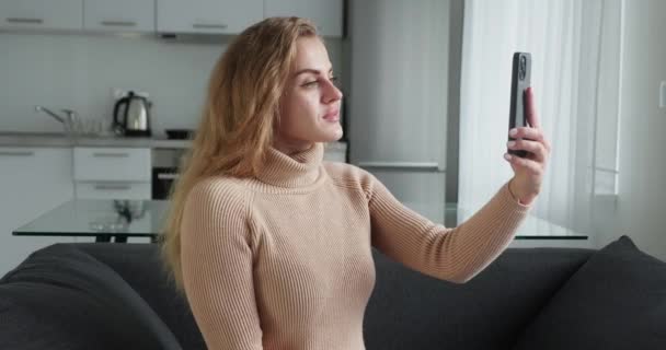 Online conference from home on kitchen background. Young pretty blonde woman of Caucasian appearance in a woolen sweater with is talking on video link using mobile phone and is emotionally happy — Stockvideo