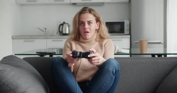 Beautiful excited young gamer woman sitting on a couch, playing and winning in video games on sofa. She plays with a wireless controller. Cozy couch in day time — Vídeo de Stock