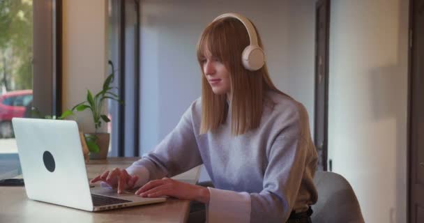 Joven alegre en los auriculares escuchando música usando el ordenador portátil en la cafetería . — Vídeo de stock