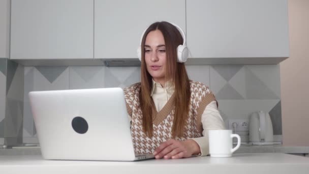 Happy caucasian young woman wear headset communicating by conference call speak looking at computer at home office, video chat job interview or distance language course class with online teacher — Stockvideo