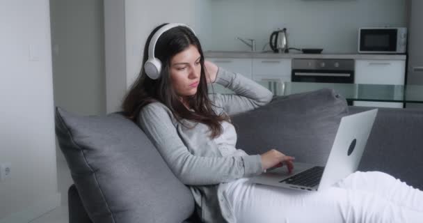 Konzentrierte junge erwachsene Frau mit Kopfhörern, die im gemütlichen Wohnzimmer auf der Couch sitzt und ihren Laptop hält. Gelegenheitsdame plaudert mit Freunden, arbeitet oder studiert von zu Hause aus online am Computer — Stockvideo