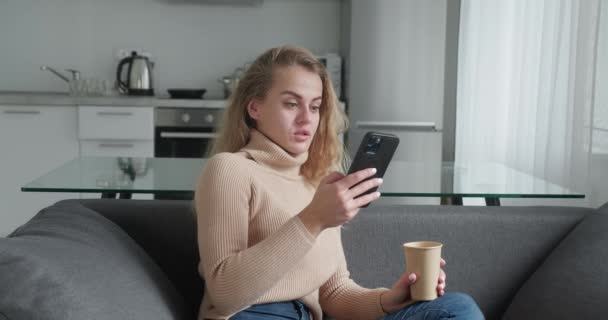 Young blonde woman at home, smiling girl using app and browsing on the internet, typing message on smartphone sitting on cozy sofa, drinking cofee, beautiful lady having pleasant funny conversation — ストック動画