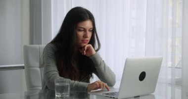 Uzun saçlı esmer kadın, mutfak arka planında otururken bilgisayarında görüntülü sohbet yapıyor. Kadın, ekrana bakarken elini sallayarak gülümsüyor. Uzaklık eğitimini kabul et.
