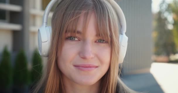 Portrét atraktivní mladé ženy s bezdrátovými sluchátky dívající se do kamery a usmívající se na pozadí města při západu slunce. Zavřít — Stock video