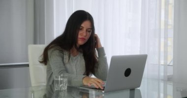 Evde dizüstü bilgisayarla çalışan serbest çalışan bir kadın. Gri kazaklı beyaz bir kız evde, masada otururken metni yazmaya, su içmeye başlıyor. Yakın plan.