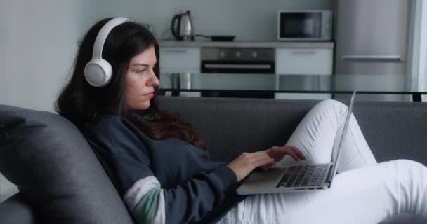 Serio bastante joven freelancer mujer trabaja en freelance desde casa moderna escribiendo correo electrónico en el ordenador portátil centrado chica uso de la computadora para el estudio en línea sentado en el sofá usuario femenino ocupado en el trabajo de Internet a distancia — Vídeos de Stock