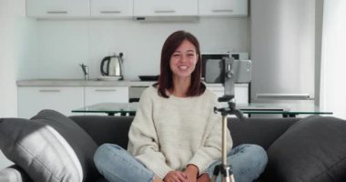 Kanepede oturan kadın yeni video günlüğü kaydediyor. Akıllı telefonu tripoda koyuyor.